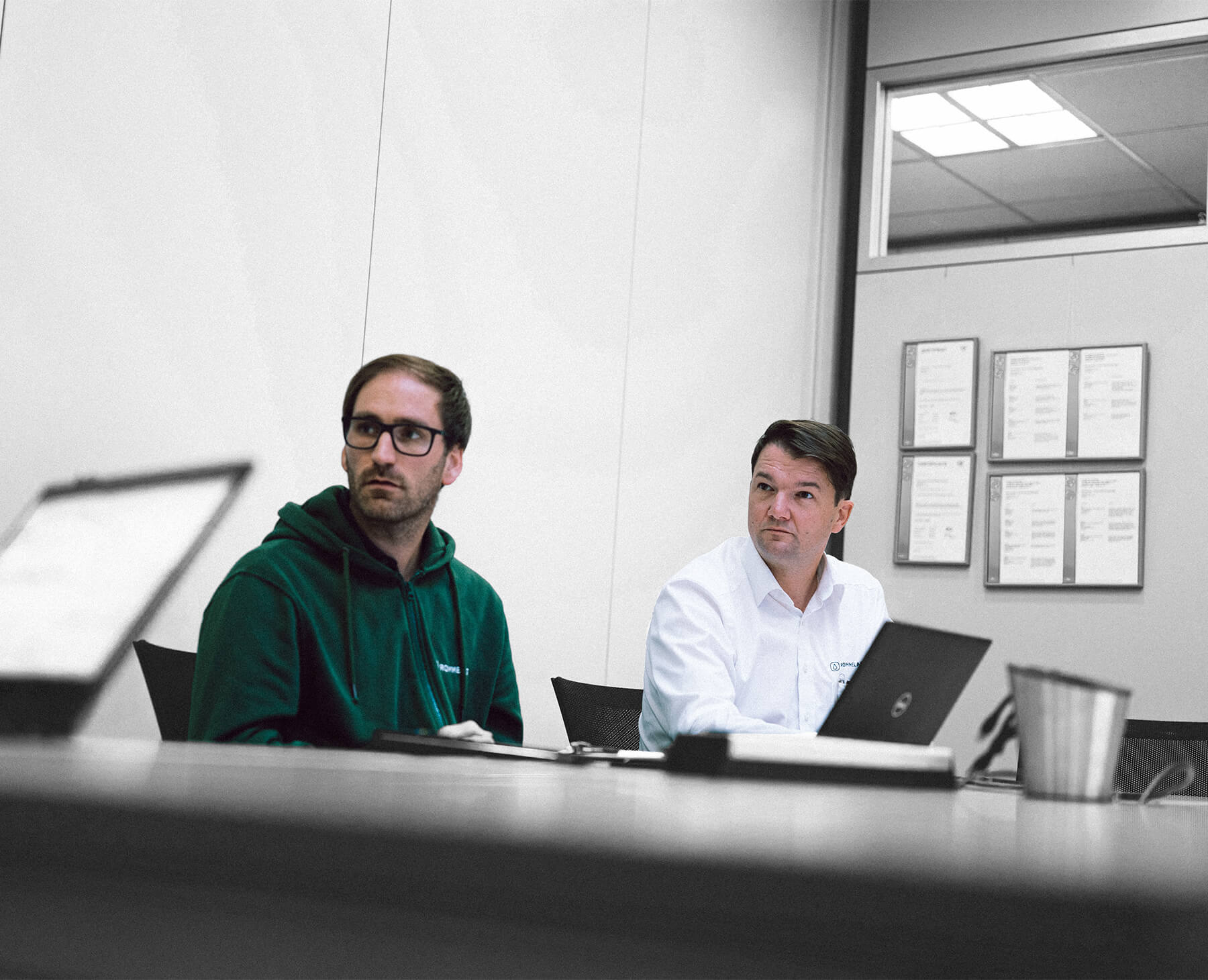 Photographie de deux hommes qui, lors d'une réunion, suivent une troisième personne non visible avec une expression faciale neutre.