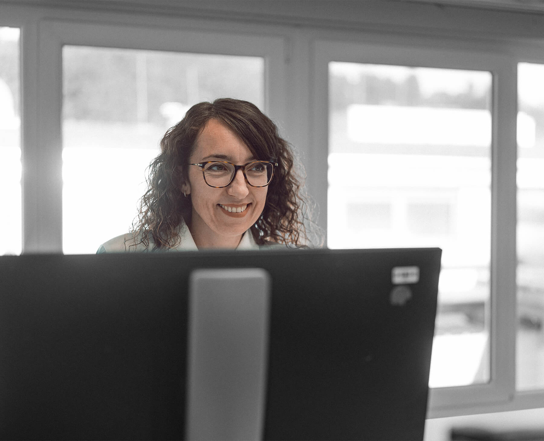 Fotografía de una mujer morena con gafas en una oficina sonriendo sobre el monitor de un ordenador.