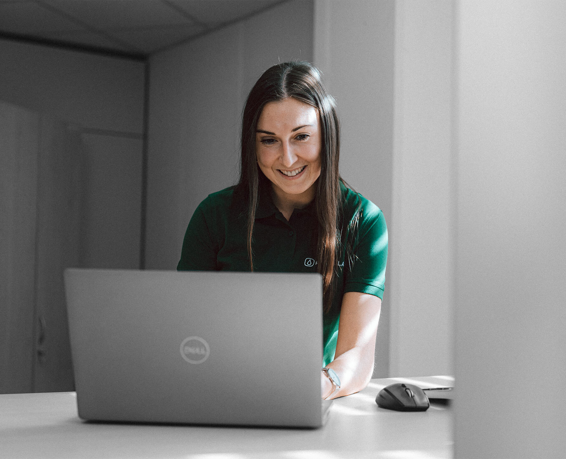 一位微笑的黑发女士在桌上摆着一台笔记本电脑,穿着一件绿色的 Rommelag T 恤,这表明联系该公司是多么容易。